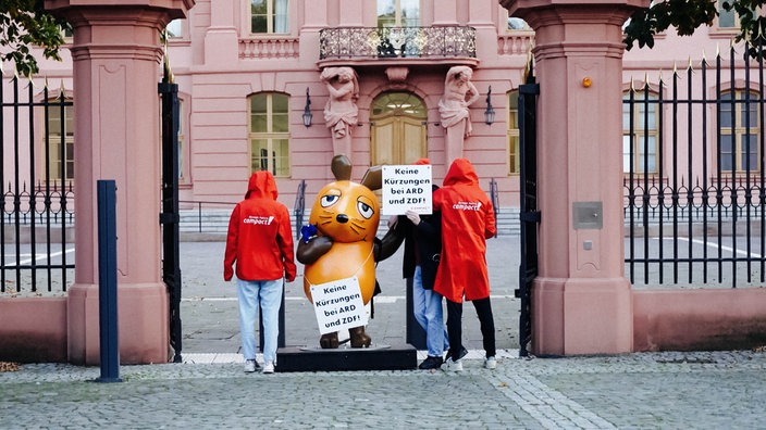 Drei Personen stehen mit der Maus-Figur im Tor des Landtags in Mainz. Sie zeigen ihre Gesichter nicht und tragen teils Campact-Jacken.