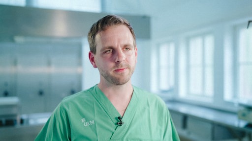 Dr. Maximilian Hagen in seiner Arbeitskleidung. Er befindet sich in einem Untersuchungszimmer.
