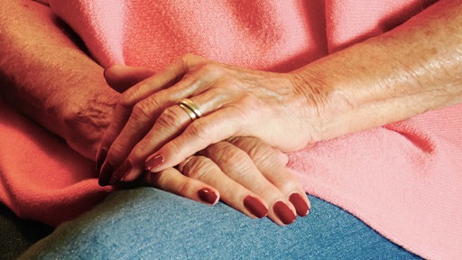 Zwei Hände liegen aufeinander. Die Nägel sind rot lackiert. Am linken Ringfinger trägt sie einen Ring.