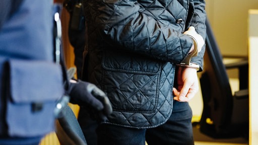 Ein Mann steht mit schwarzer Jacke und schwarzer Hose, die Hände in Handschellen, neben einem Polizisten, von beiden sieht man nur die Körpermitte