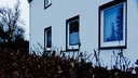 Fassade eines weiß gestrichenen Hauses mit drei sichtbaren Fenstern, davor ragt eine Hecke