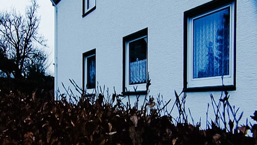 Fassade eines weiß gestrichenen Hauses mit drei sichtbaren Fenstern, davor ragt eine Hecke