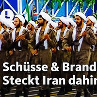 Iranische Revolutionsgarden marschieren mit Gewehren über eine feierlich geschmückte Straße. Dazu steht folgender Text: Schüsse und Brandanschlag - Steckt Iran dahinter?