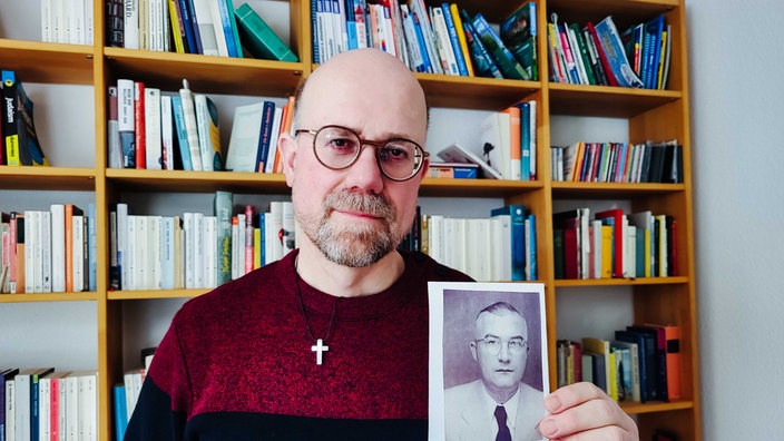 Ein Mann steht vor einem Bücherregeal, er trägt einen rot-schwarzen Pullover, eine Brille, Dreitagebart und eine Glatze, in der Hand hält er ein schwarz-weiß Foto seines Opas 