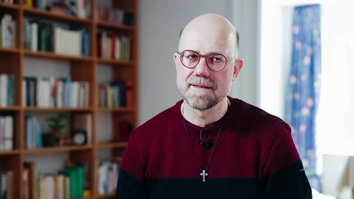 Mann mit Brille, Dreitagebart und Glatze, rot-schwarzes Hemd, darüber eine Kreuzkette, links ein Bücherregal