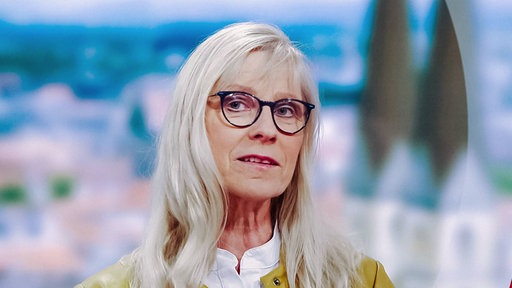 Frau mit weißen Haaren, schwarzer Brille, weißer Bluse und gelber Jacke, Mund leicht geöffnet, Blick nach links