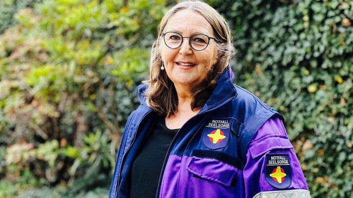 Therese Mauer steht vor einem grünen, waldigen Hintergrund. Sie trägt eine runde Brille und eine Einsatzjacke der Notfallseelsorge.