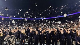 Eine Arena voller Kommissarsanwärter. Sie alle schmeißen ihre Mützen in die Luft.