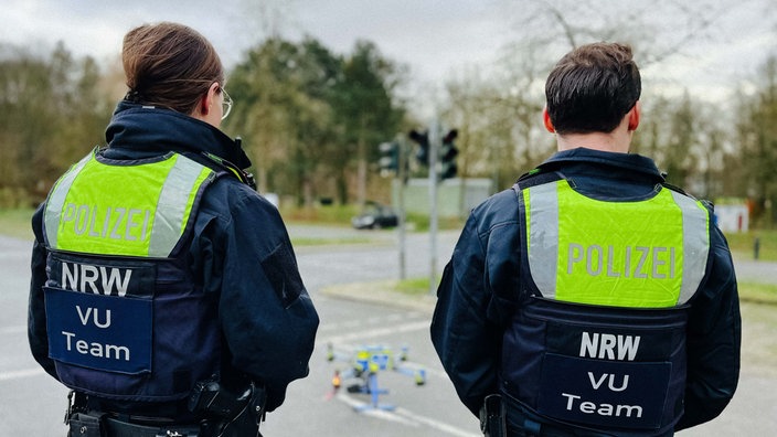 Zwei Polizisten stehen mit dem Rücken zur Kamera. Sie tragen jeweils eine Weste mit der Aufschrift "Polizei NRW VU Team".