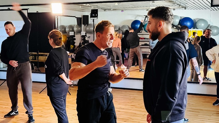 Trainer Dirk Eisenblätter mit erhobener Faust einem Kursteilnehmenden gegenüber. Im Hintergrund weitere Kursteilnehmende.