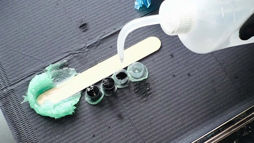 Auf einer grauen Matte liegen verschiedene Tätowierwerkzeuge: Farbe in kleinen Töpfen, ein Holzspatel, ein grünes Gel und eine große Farbdose.