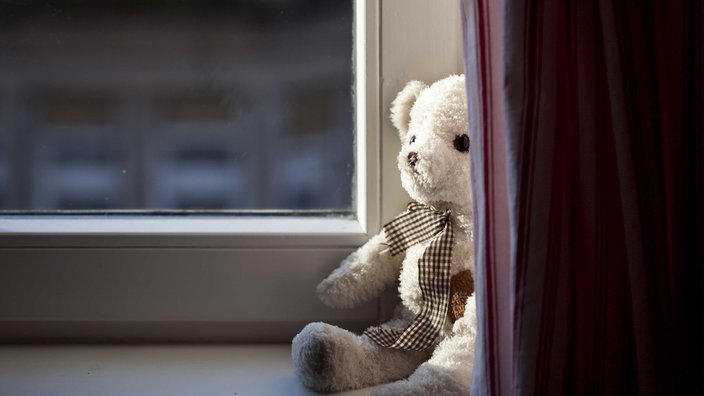 Ein weißer Teddybär sitzt auf einer Fensterbank, zur Hälfte von einem Vorhang verdeckt