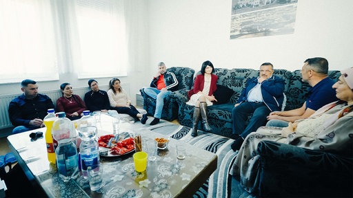 Mehrere Erwachsene sitzen in einem Raum auf einem Sofa und auf dem Boden verteilt. 