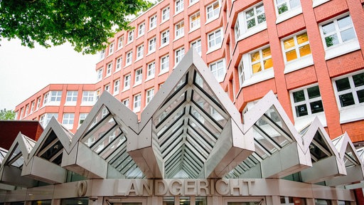 Ein gezacktes Vordach aus Metall und Glas, darunter der Schriftzug "Landgericht" über einer Schiebetür. Im Hintergrund ragt ein mehrstöckiges Bürohaus aus rotem Backstein hervor.