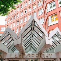 Ein gezacktes Vordach aus Metall und Glas, darunter der Schriftzug "Landgericht" über einer Schiebetür. Im Hintergrund ragt ein mehrstöckiges Bürohaus aus rotem Backstein hervor.