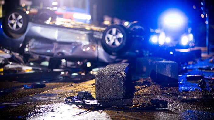 Ein schwerer Autounfall nachts. Ein Auto liegt zerbeult auf dem Dach. Im Hintergund sieht man das Blaulicht eines Krankenwagens.
