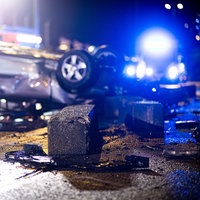 Ein schwerer Autounfall nachts. Ein Auto liegt zerbeult auf dem Dach. Im Hintergund sieht man das Blaulicht eines Krankenwagens.