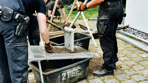 Drei Männer graben in einem Garten nach Knochen. Zwei von ihnen sieben die Erde.