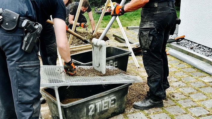 Drei Männer graben in einem Garten nach Knochen. Zwei von ihnen sieben die Erde.