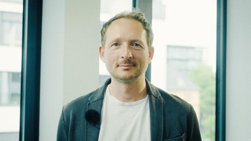 Ein Mann mit weißem T-Shirt und grauer Jacke steht in einem Büro, hinter ihm ein Fenster.