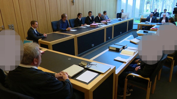 Mehrere Personen sitzen in einem Gerichtssaal des Landgerichts Münster. Die Gesichter von drei Personen sind gepixelt.
