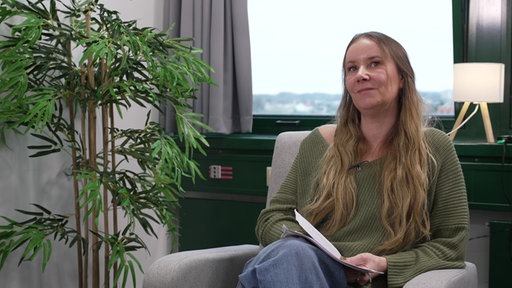 Kriminaloberkommissarin Maike Schmidt sitzt auf einem Stuhl vor einem Fenster und hält eine Akte in den Händen.