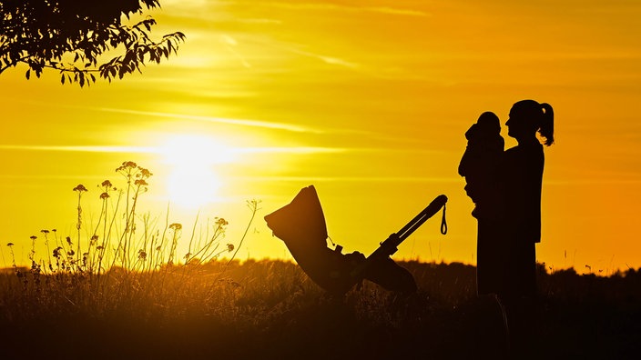 Symbolbild: Eine Frau hält ein kleines Kind auf dem Arm steht auf einem Feldweg im gelben Gegenlicht eines Sonnenuntergangs, daneben ein Kinderwagen