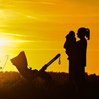Symbolbild: Eine Frau hält ein kleines Kind auf dem Arm steht auf einem Feldweg im gelben Gegenlicht eines Sonnenuntergangs, daneben ein Kinderwagen