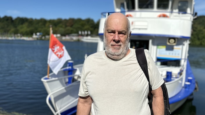 Ein Mann in braunem T-Shirt steht vor einem blau-weißen Boot, was ankert. Der Himmel ist blau und die Sonne scheint. 
