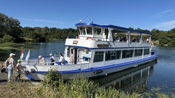 Ein blau-weißes Boot ankert an einem pflanzenbewachsenen Ufer, Menschen steigen ein und aus. 