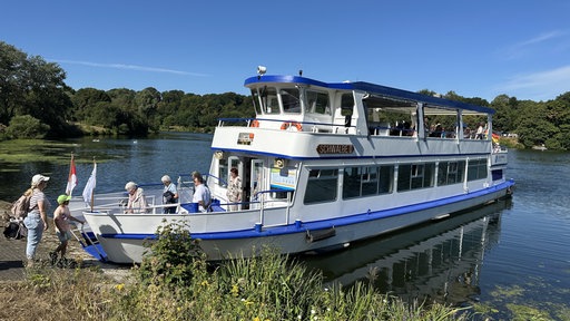 Ein blau-weißes Boot ankert an einem pflanzenbewachsenen Ufer, Menschen steigen ein und aus. 