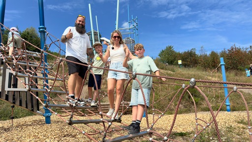 Ein Mann, eine Frau und zwei Jungen stehen auf einem Spielplatz und lächeln in die Kamera 