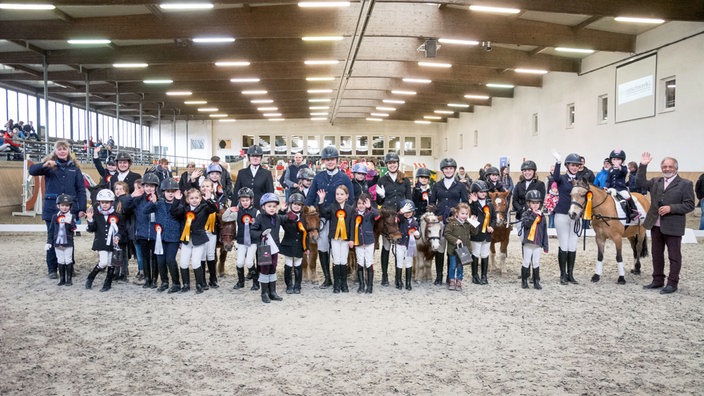 Eine große Gruppe aus Kindern und Erwachsenen in Turnierkleidung und Preisschleifen steht mit ihren Pferden in einer Reithalle.