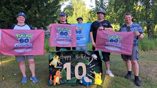 Fünf Menschen stehen mit "Pokemon GO"-Bannern hinter einem "Happy Birthday Pokemon GO"-Plakat.