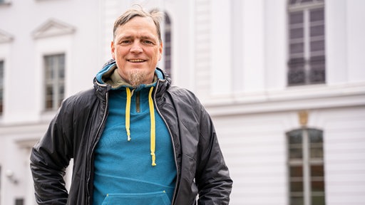 Ein Mann mit Outdoor-Kleidung steht vor einem Universitätsgebäude.