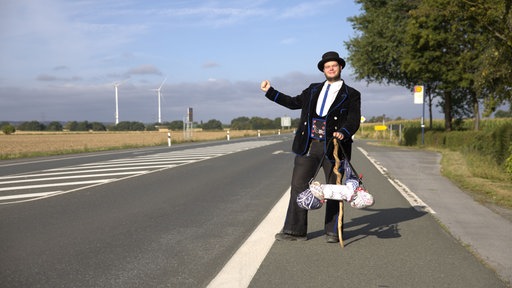 Handwerker Oliver Pesse auf der Walz steht neben einer Landstraße bei Soest, während ein Auto vorbeifährt