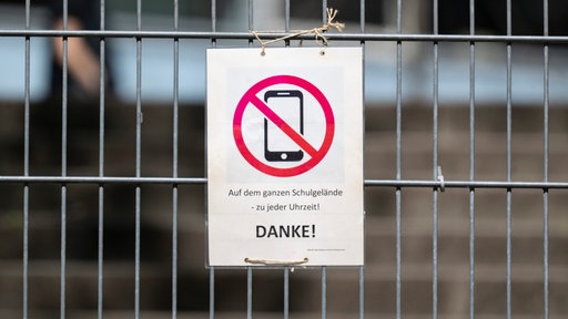 Ein Schild mit einem durchgestrichenen Smartphone und der Aufschrift «Auf dem ganzen Schulgelände - zu jeder Uhrzeit! DANKE!» hängt am Zugangstor zum Schulhof einer Schule
