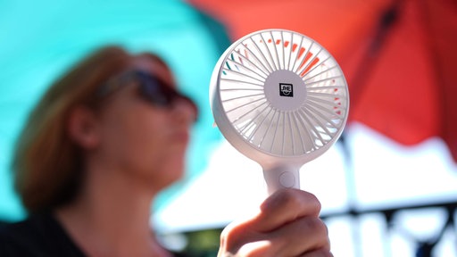 Eine Frau kühlt sich mit einem Handventilator unter Sonnenschirmen auf einem Balkon in der Mittagssonne ab