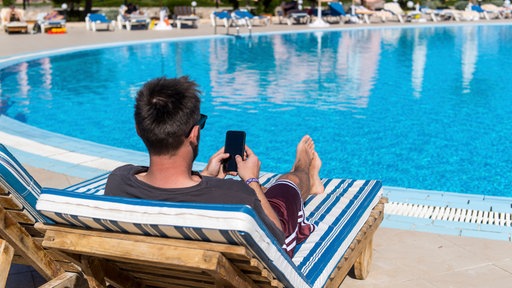 Ein Mann liegt an einem Hotelpool auf einer Sonnenliege und schaut auf sein Smartphone
