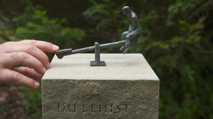 Ein Finger drückt eine Messingfigur auf einer Wippe in die Luft