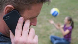  Ein Vater sitzt auf einer Gartenliege und telefoniert mit einem Smartphone, während im Hintergrund ein Mädchen alleine mit einem Ball im Garten spielt.