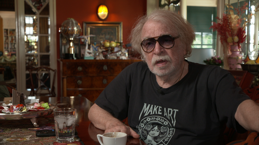 Zirkusdirektor Bernhard Paul trägt eine große Sonnenbrille und sitzt seitlich am Tisch.