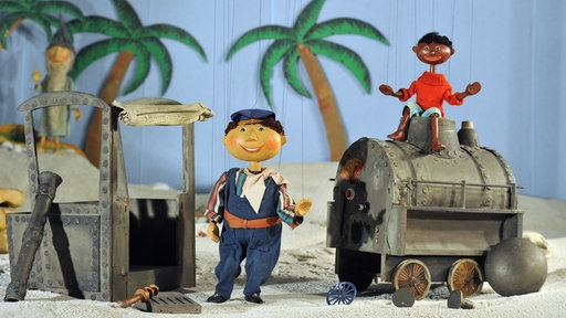 Die Marionetten Scheinriese Tur Tur (hinten), Lukas und Jim Knopf im Puppenkistenmuseum mit der kaputten Lokomotive Emma in einem Wüstenszenario