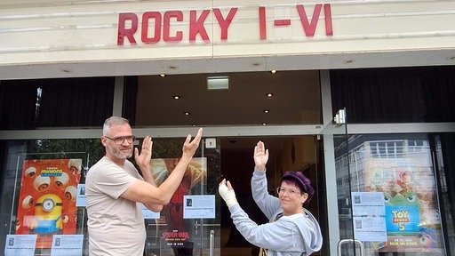 Ein Mann und eine Frau zeigen auf den Schriftzug "Rocky 1-6" über dem Eingang eines Kinos.