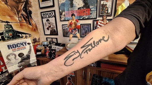 Ein Unterarm mit der Unterschrift von Sylvester Stallone, dahinter Rocky-Fanmerchandise.