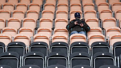 Symbolbild: Ein Fan sitzt auf einer Tribüne im Fußballstadion, deren Plätze ansonsten alle leer sind und liest in einem Buch.