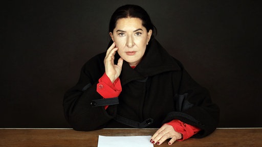 Die Künstlerin Marina Abramović sitzt an einem Schreibtisch und guckt direkt in die Kamera