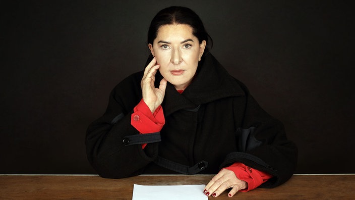 Die Künstlerin Marina Abramović sitzt an einem Schreibtisch und guckt direkt in die Kamera