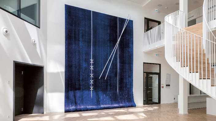 Ein blauer Wandteppich hängt an der Wand eines hellen Atriums.