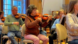 Eine ältere Frau sitzt in einem Orchester und spielt die Geige.
